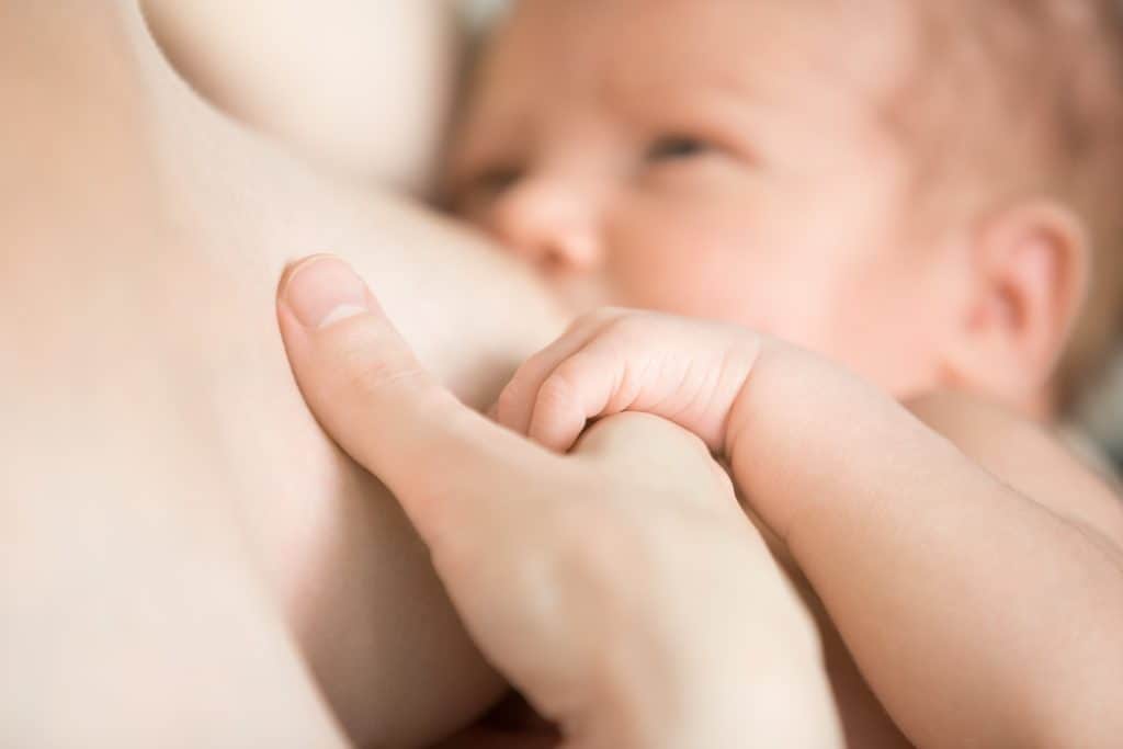 Close-up portrait of sweet newborn baby breastfeeding. Adorable new born caucaian kid holding moms finger with his cute little palm while breast feeding. Focus on hands. Young mother nursing baby